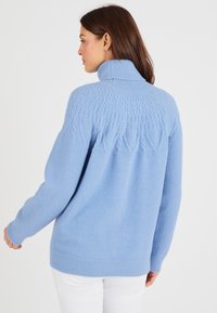 Un pull en tricot bleu clair avec un col haut, présentant des motifs texturés sur les épaules et le dos, associé à un pantalon blanc.