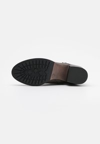 Suela de bota de cuero marrón con patrón de tracción de goma negra y tacón bajo, apoyada sobre un fondo blanco.