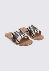 Chanclas planas con suela de cuero marrón que presentan un patrón a rayas en negro y crema en la parte superior, junto con acentos en cuero negro.