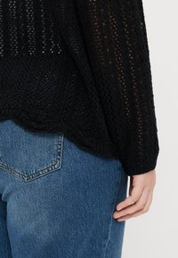 Black knitted sweater with a loose, open weave design, featuring a scalloped hem and paired with blue denim jeans.