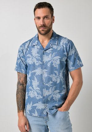 Man met baard en getatoeëerde arm draagt een blauw bloesje met korte mouwen en knopen, met hand in de zak van lichtblauwe spijkerbroek.