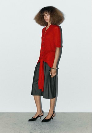 Camicia rossa a maniche corte, annodata in vita, abbinata a culotte grigie ampie e decolletè neri a punta con cinturino.