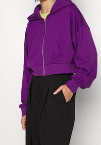 Hoodie cropped viola con zip frontale e grande tasca a scomparsa. Presenta maniche ampie e polsini a coste. Abbinato a pantaloni neri a vita alta.