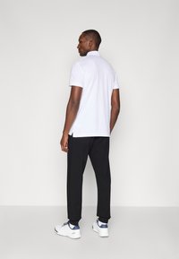 Vit polo shirt med ett texturerat mönster, svarta sweatsbyxor med mudd och vita träningsskor med blå detaljer, sedd bakifrån.