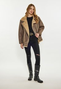 Giacca in shearling marrone con colletto ampio, abbinata a jeans neri strappati e stivali neri in pelle al ginocchio.