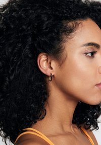 Złoty i czarny kolczyk w kształcie koła noszony w lewym uchu. Kolczyk charakteryzuje się gładkim, okrągłym wzorem z kontrastowym detalem kolorystycznym.