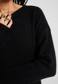 Pull en laine noire à coupe ample, col large et tissu texturé. Comprend des manches longues et des doigts anneaux visibles tenant le tissu.