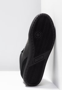 Semelle de basket noire à motif ondulé appuyée contre un coin blanc, montrant le nom de la marque et un logo rond près du talon.