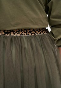 Spódnica tiulowa w oliwkowej zieleni z paskiem w leoparda. Przezroczysty, lekki materiał tworzy warstwowy wygląd.