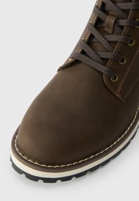 Brown leather boot featuring a round toe, sturdy laces, gold eyelets, and a contrasting white edge on the sole with a textured black tread.