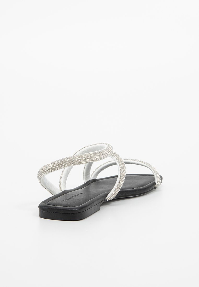 Vlakke zwarte sandalen met witte banden versierd met kleine zilveren kralen. Gladde textuur en minimalistisch ontwerp zonder omgebogen teen of hak.