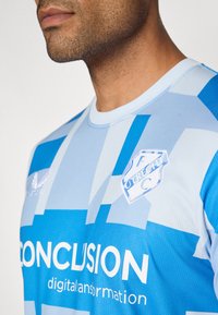 Modra športna majica z geometrijskimi svetlobnimi in temno modrimi vzorci. Na sprednji strani vključuje logo in besedilo v beli barvi. Narejena je iz zračnega materiala.