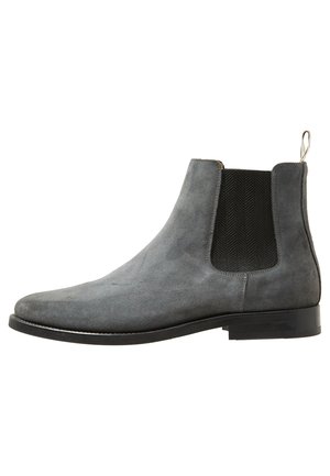 Bottine Chelsea en daim gris avec panneau latéral élastique noir et semelle noire, avec une languette à l’arrière.