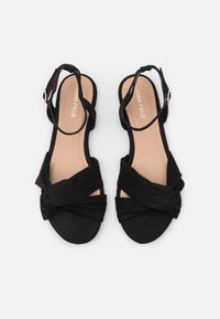 Paire de sandales plates noires avec des brides avant torsadées et des boucles à la cheville, semelles intérieures beiges étiquetées "ANNA FIELD,", présentées sur fond blanc.