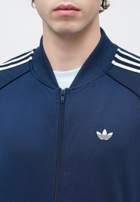 Μπλε ναυτικού χρώματος ζακέτα με φερμουάρ, κατασκευασμένη από υφή υφάσματος, με λευκές τριπλές ρίγες στους ώμους και μικρό λευκό λογότυπο στο στήθος.