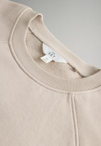 Beige sweatshirt met geribbeld kraag en manchetten, voorzien van een zachte textuur en een genaaid label met de tekst "THE SET" in het zwart. Maat M aangegeven.