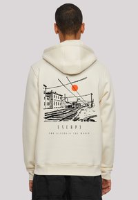 Person wearing cream hoodie with black sketch of train tracks, buildings, orange sun, and text "ESCAPE and discover the world" on the back.