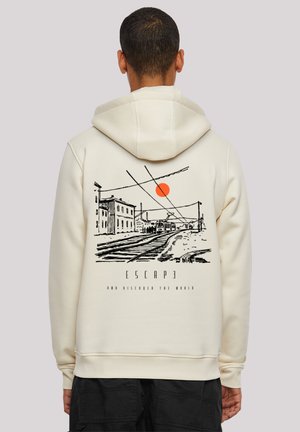 Person wearing cream hoodie with black sketch of train tracks, buildings, orange sun, and text "ESCAPE and discover the world" on the back.