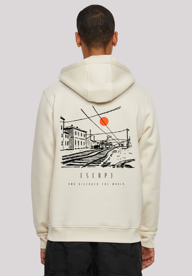 Person wearing cream hoodie with black sketch of train tracks, buildings, orange sun, and text "ESCAPE and discover the world" on the back.
