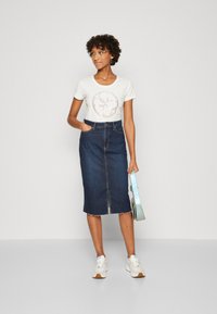 Camiseta blanca con gráfico circular, falda vaquera lápiz con abertura frontal, zapatillas deportivas y un bolso liviano azul. Conjunto casual con texturas variadas.