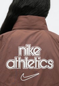 Καφέ ελαφρύ μπουφάν με ψηλό γιακά, λευκό λογότυπο "nike athletics" και λεπτομέρεια Nike swoosh στην πλάτη. Λεπτή υφή.