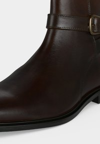 Bottine en cuir marron foncé avec une bride décorative et une boucle en laiton, dotée d’un petit talon noir et d’une finition lisse.