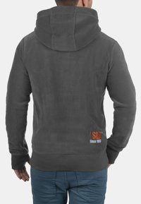 Gray hoodie with a soft fleece texture, featuring a large hood, ribbed cuffs, and an embroidered logo in orange and white at the lower back.