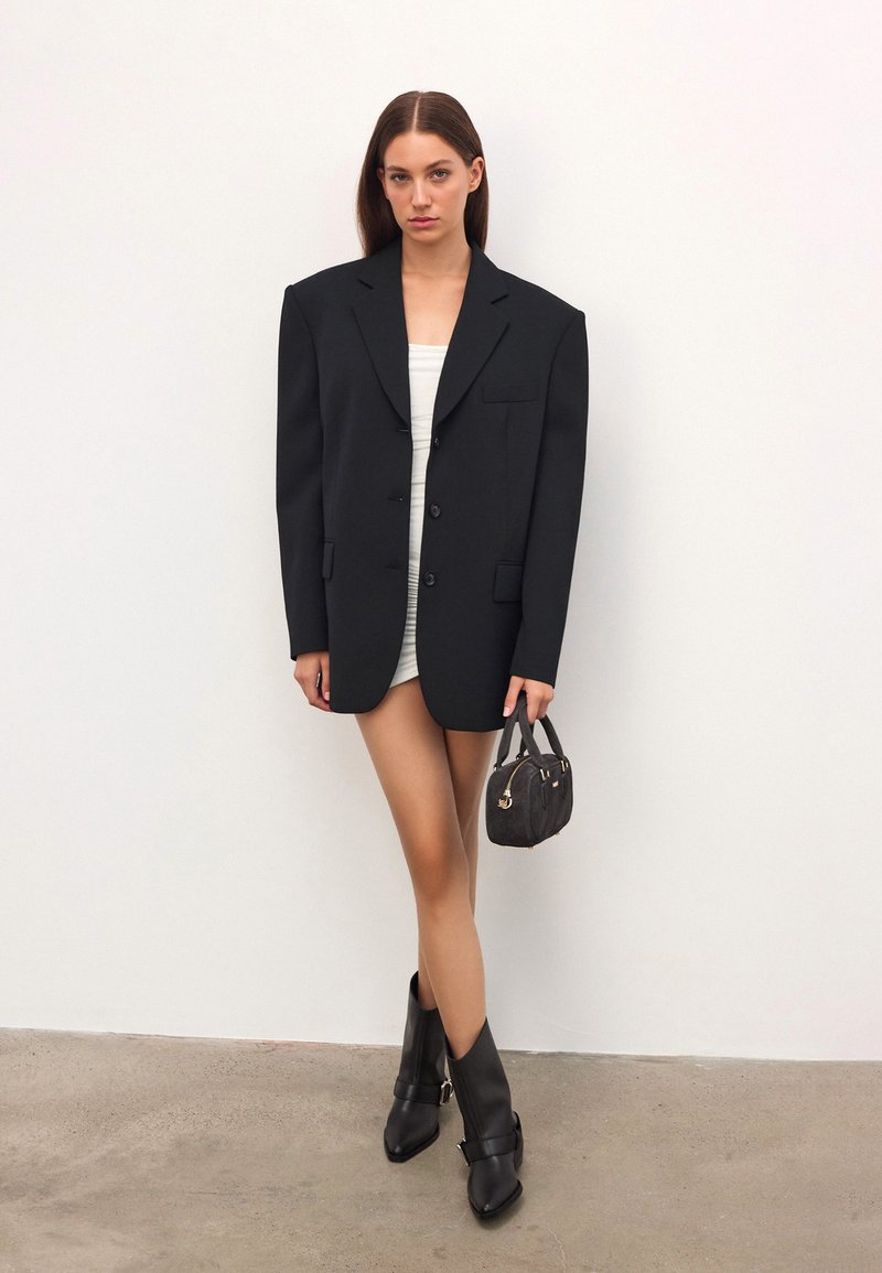 Overmaat zwarte blazer met gesneden revers, enkele borstzak en twee knopen, gedragen over een goed passend witte jurk. Zwarte laarzen met hak en tas.