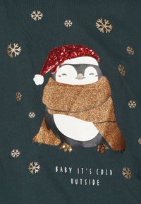 Camiseta verde con un pingüino que lleva un sombrero rojo brillante y una bufanda dorada brillante, rodeado de copos de nieve y el texto: "HACE FRÍO AFUERA."