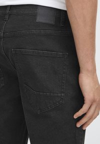 Blugi negri din denim cu textură fină, două buzunare în spate și o etichetă simplă de piele la talie. Vedere de aproape din lateral.