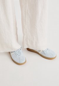 Baskets en daim bleu clair avec un accent crème, dotées d'un design à lacets et de semelles en caoutchouc, associées à un pantalon large de couleur claire.