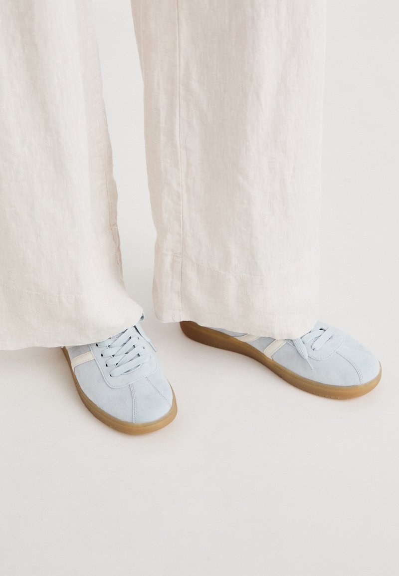 Baskets en daim bleu clair avec un accent crème, dotées d'un design à lacets et de semelles en caoutchouc, associées à un pantalon large de couleur claire.
