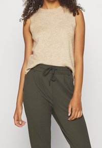 Donna che indossa un top in maglia senza maniche beige e pantaloni verde oliva con coulisse, in piedi con le braccia rilassate su uno sfondo chiaro e semplice.