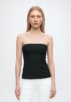 Donna con capelli biondi lisci che indossa orecchini a cerchio dorati, un top nero senza spalline e pantaloni bianchi con ricamo floreale.