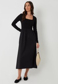 Vestido negro con escote cuadrado, mangas largas y falda fruncida. Combinado con bailarinas negras y un bolso beige. Textura de tela suave.