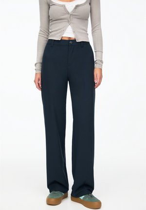 Femme portant un pantalon large foncé, un cardigan beige court boutonné sur un haut blanc, et des baskets grises avec des semelles marron.