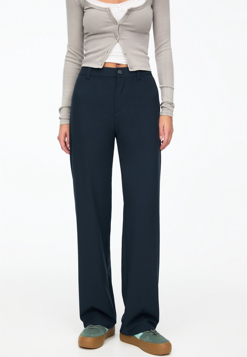 Femme portant un pantalon large foncé, un cardigan beige court boutonné sur un haut blanc, et des baskets grises avec des semelles marron.