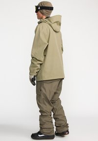 Khaki skijas met capuchon, gecombineerd met bijpassende cargobroek. Kenmerken zijn handschoenen en skibril op een neutrale achtergrond.
