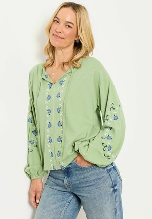 Woman with blonde hair wearing a light green blouse with blue floral embroidery and blue jeans, smiling with one hand in pocket.