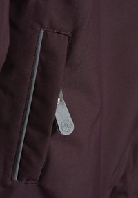 Burgundy ytterkläder med en ficka som stängs med dragkedja och en grå dragflik. Snyggt, slitstarkt tyg med kontrasterande grå sömmar och detaljer.