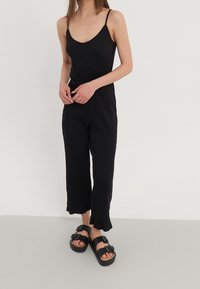 Combinaison noire côtelée avec un décolleté en V, des bretelles fines et des jambes larges et raccourcies, assortie à des sandales noires à double boucle.