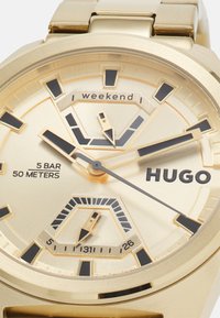 Reloj en tono dorado con un dial texturizado, que presenta acentos negros, tres sub-diales y el nombre de la marca "HUGO". Resistente al agua hasta 50 metros.