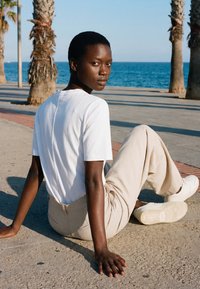 Jeune personne en chemise blanche et pantalon beige assise sur le trottoir près des palmiers au bord de la mer, regardant par-dessus son épaule vers la caméra.