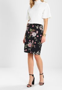 Vit topp med vida ärmar, kombinerad med en svart pennkjol med färgglada blommönster. Svarta klackar med remmar runt anklarna. Guldarmband på handleden.