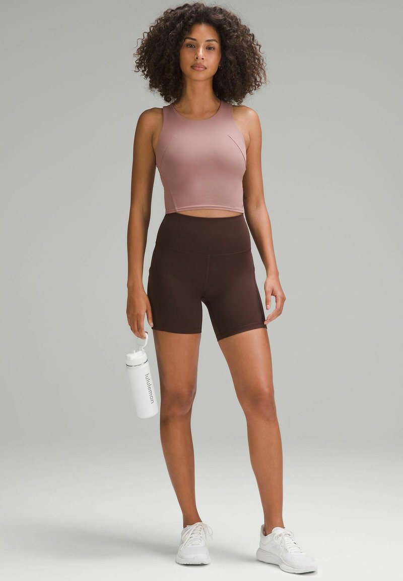 Hellrosa, kurz geschnittenes Sportoberteil und braune, hochgeschnittene Shorts; eine Frau hält eine weiße Wasserflasche und trägt weiße Sportschuhe.