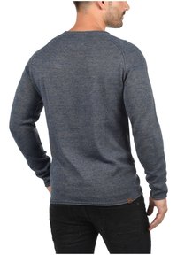 Kék kötött pulóver hosszú ujjakkal és kerek nyakkivágással. Texturált felülettel rendelkezik, és a szegélyén egy kis logós címke található.