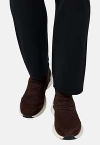 Tamsiai rudos mezginiai sportbačiai su baltais padais, dėvimi su juodomis kelnėmis, parodyti iš priekio, žiūrint iš žemesnio kampo.