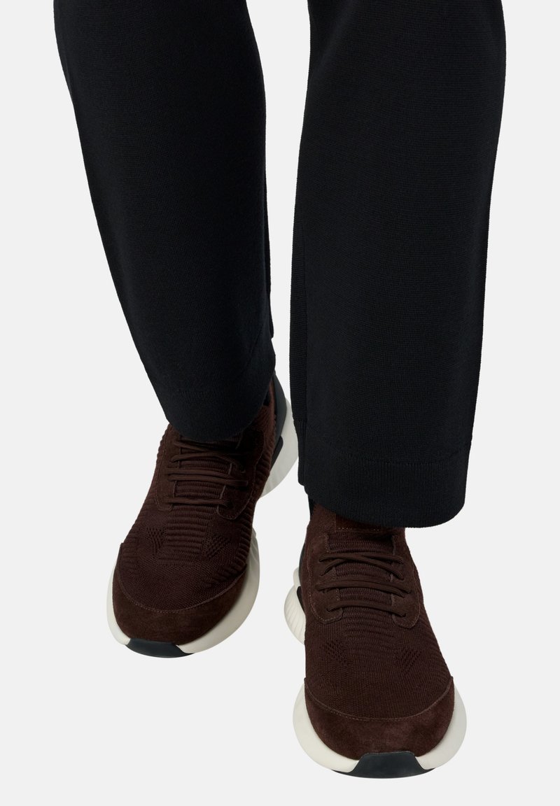 Tamsiai rudos mezginiai sportbačiai su baltais padais, dėvimi su juodomis kelnėmis, parodyti iš priekio, žiūrint iš žemesnio kampo.