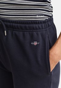 Marineblaue GANT Jogginghose mit elastischem Bund und Kordelzug, getragen mit einem schwarz-weiß gestreiften, in die Hose gesteckten Oberteil.