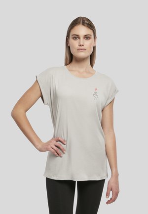 Jeune femme aux cheveux raides portant un t-shirt gris clair avec un petit motif main et cœur rouge sur la poitrine, debout avec une main sur la hanche.